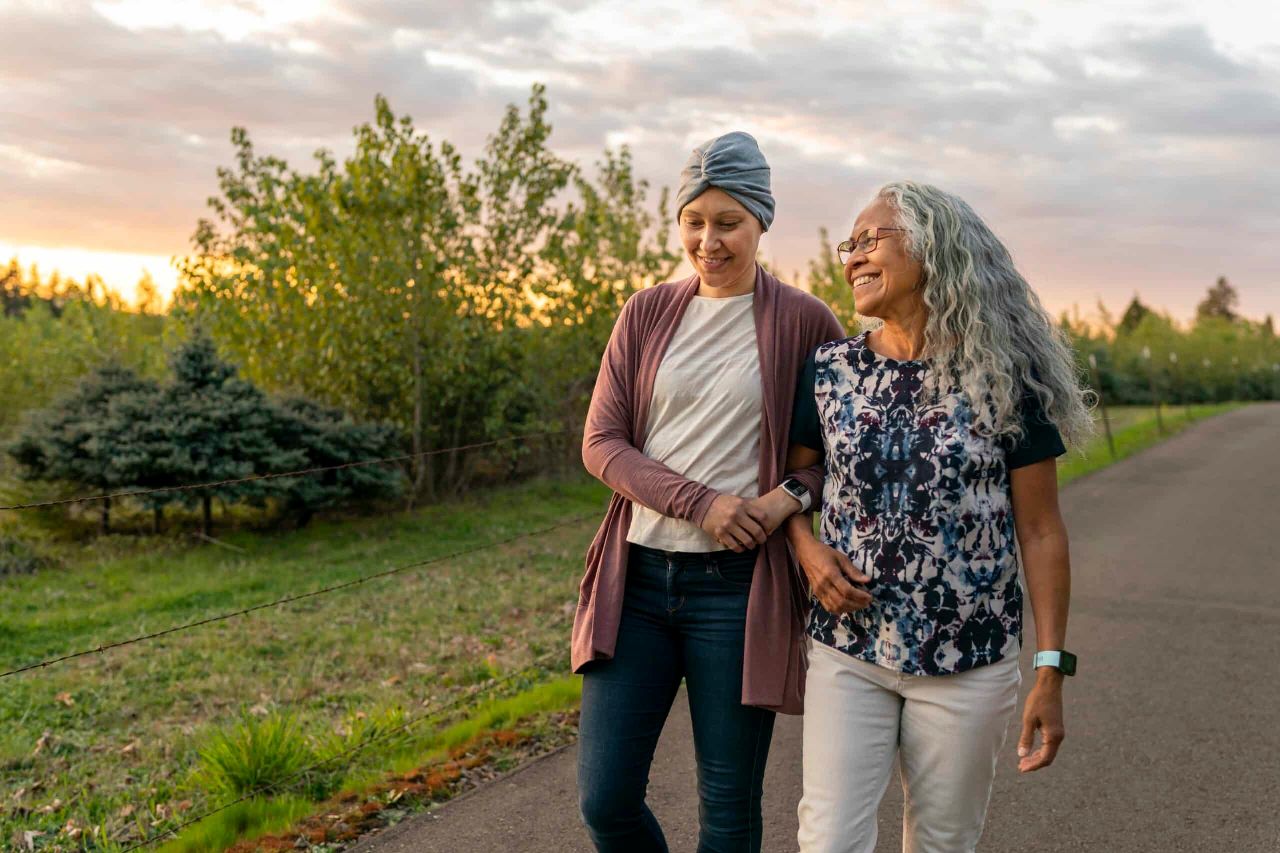 A young mixed race woman recovering from cancer walks arm in arm with her senior mother. They are enjoying the outdoors and time together. They are walking down a rural road at sunset.